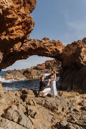 Man proposes to woman on rocky coastal arch with ocean waves and blue sky backdrop.