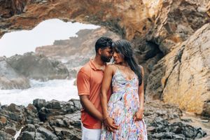 Couple sharing a romantic moment in a rocky sea cave, woman in floral dress with baby bump, waves in background.