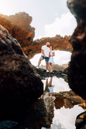 Couple standing romantically on rocky shore framed by natural stone arch with water reflection below.