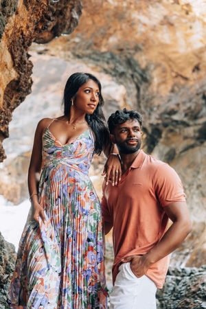 Couple posing in a rocky coastal cave, woman in floral dress and man in coral polo shirt.