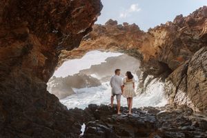 Couple standing hand in hand inside a rocky sea cave arch with crashing ocean waves in the background.