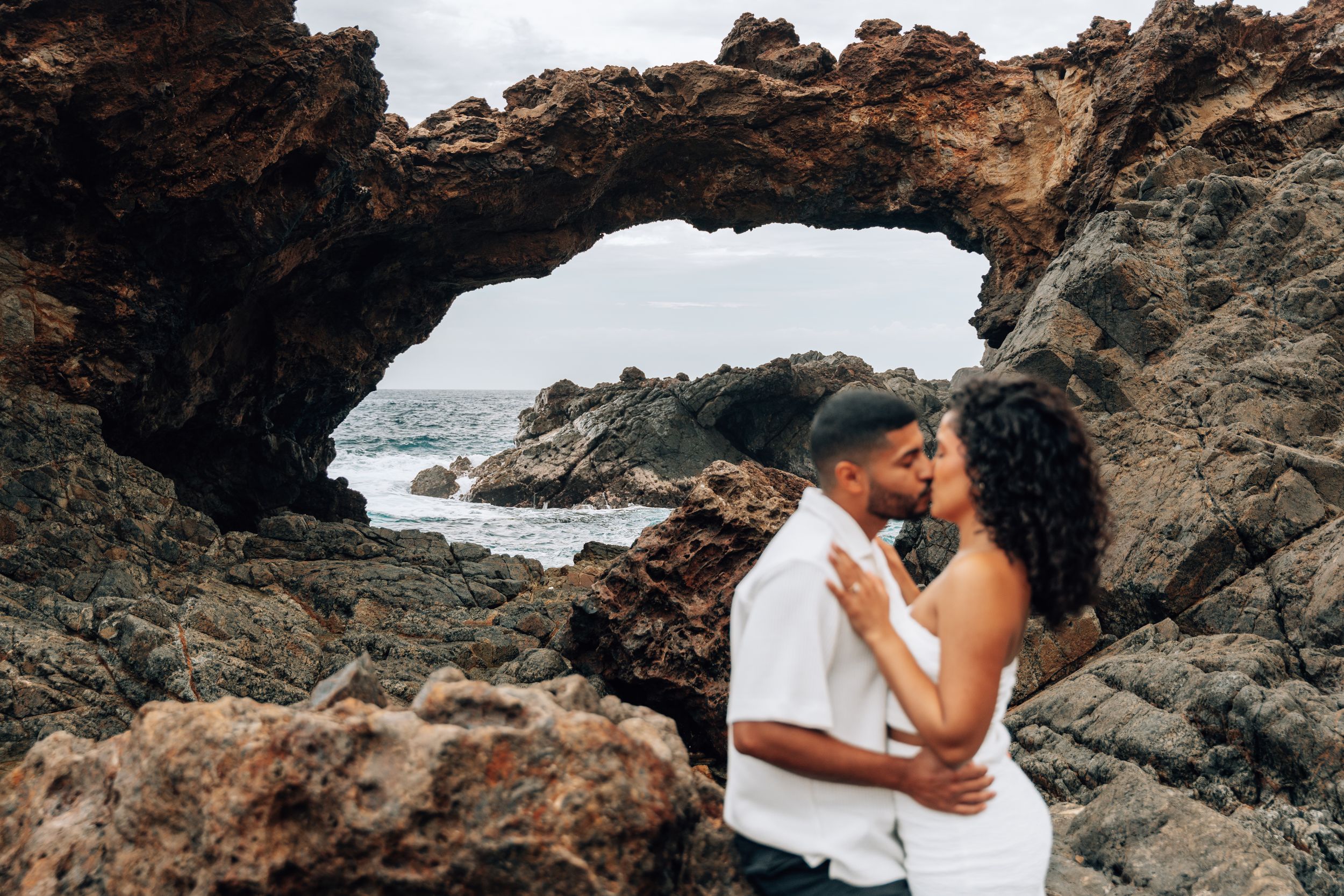 Couple sharing a romantic moment framed by a natural rock arch on a rugged coastline with ocean waves behind them.