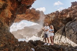 Couple standing inside a dramatic rocky arch on a rugged coastline with crashing waves behind them.