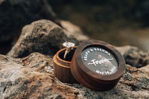 Wooden ring box engraved with "Frank & Reshma Forever May 14, 2022" holding a diamond engagement ring on rocks.