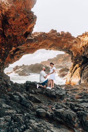 Man proposing to woman on rocky coastal arch with ocean waves, romantic outdoor engagement scene.