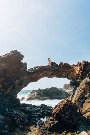 Couple embracing atop a dramatic natural rock arch over the ocean under a bright blue sky.