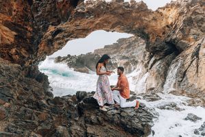 Man proposing to woman on rocky shore inside a natural cave arch with ocean waves crashing behind them.