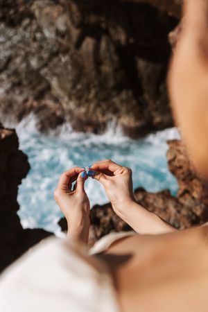 Person holding a blue gemstone ring with rocky coastal cliffs and turquoise ocean waves in the background.