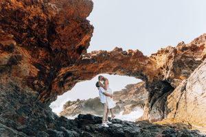 Couple embracing under a natural rock arch on a rugged coastline with ocean waves in the background.