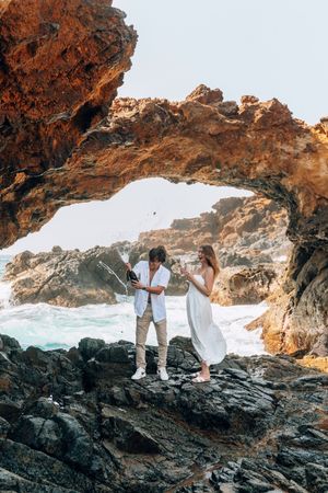 Couple celebrating with champagne under a natural rock arch by the ocean waves.