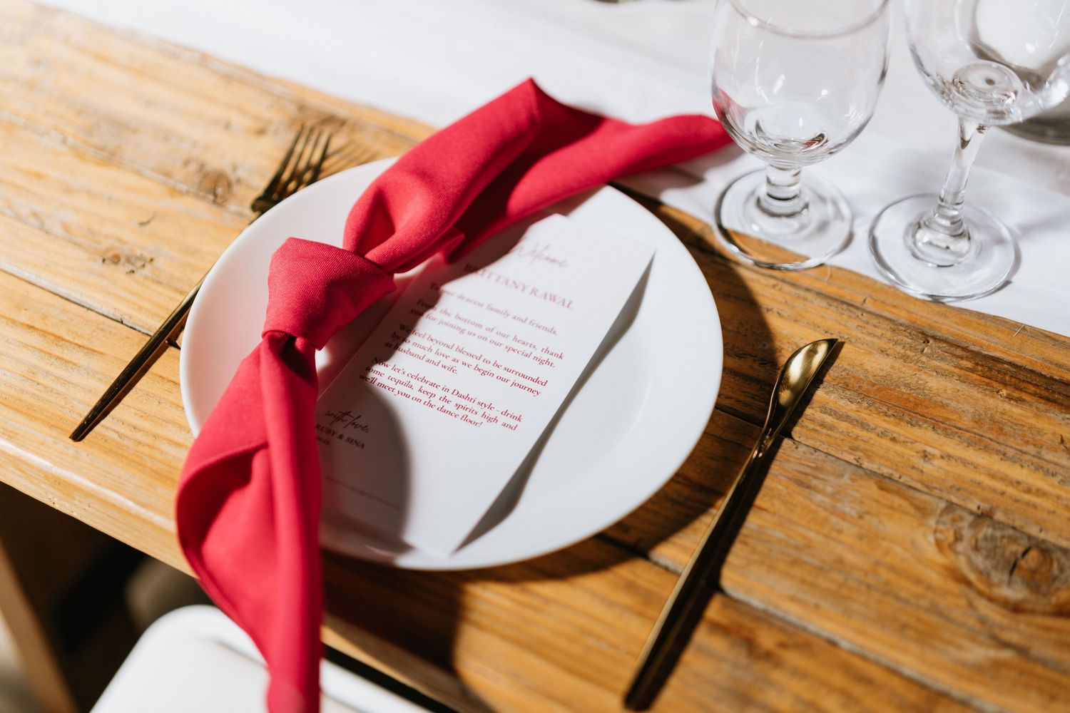 Wedding reception place setting with a red napkin, printed menu, glassware, and gold flatware.