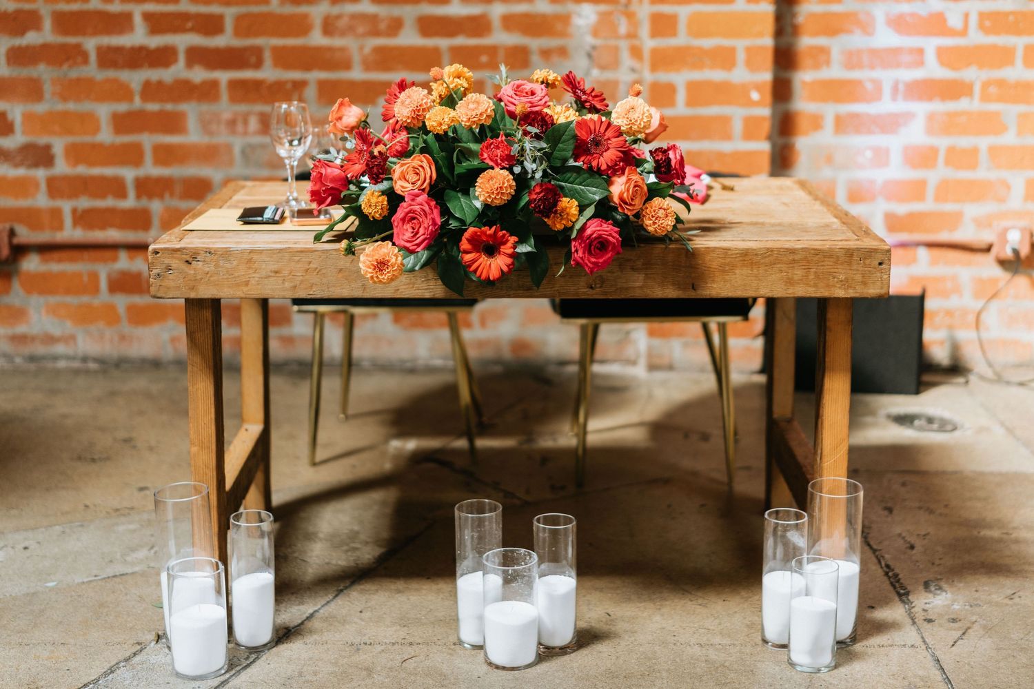 Sweetheart table decorated with a vibrant floral arrangement and glass candles against a brick wall.