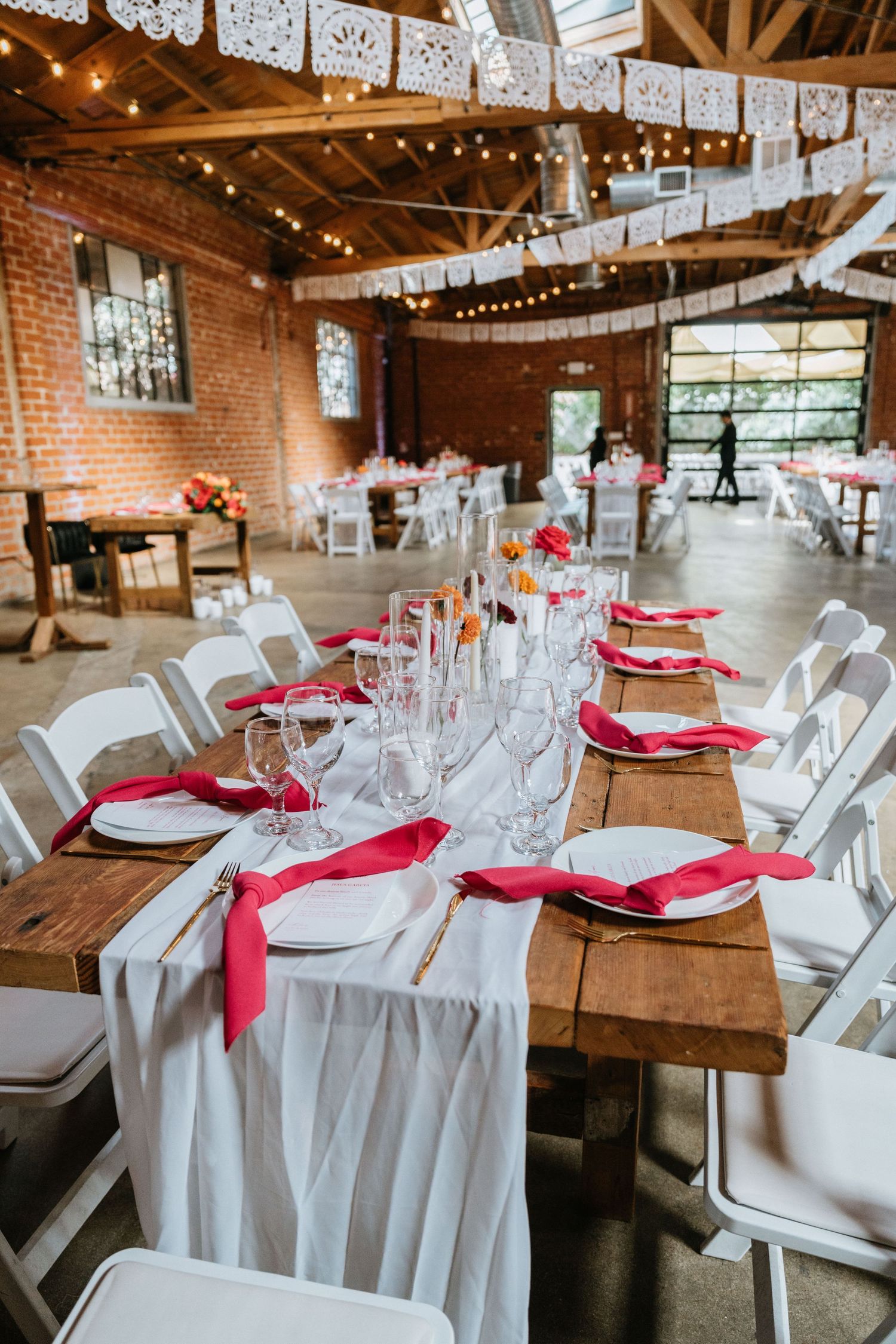 Long wooden reception table set with white plates, red napkins, candles, and glassware.