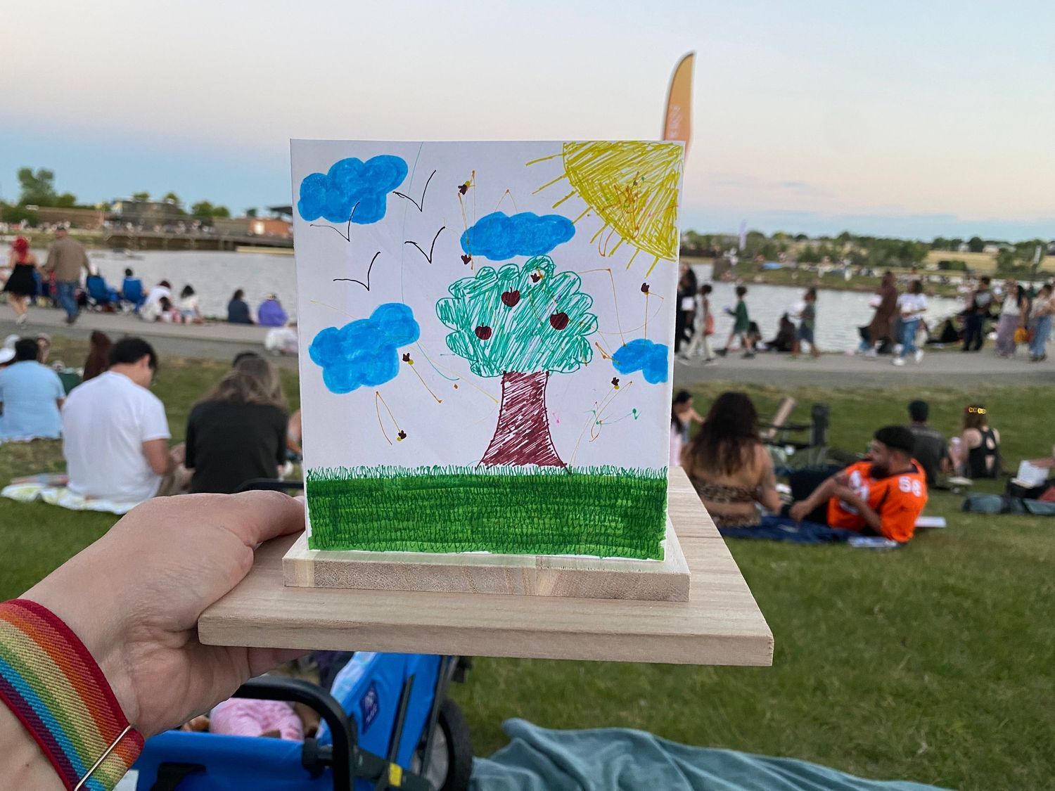 Hand holding a child's colorful drawing of a tree with clouds and sun on a wooden easel at an outdoor park event.