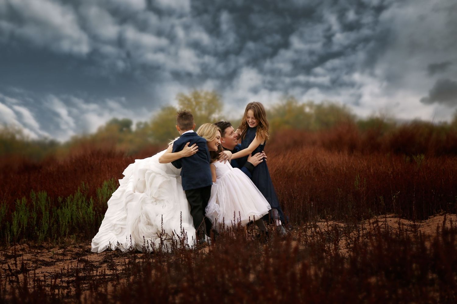 A family embraces in a red-grass field under a dramatic stormy sky, with two girls in white tulle gowns and a boy in a suit.