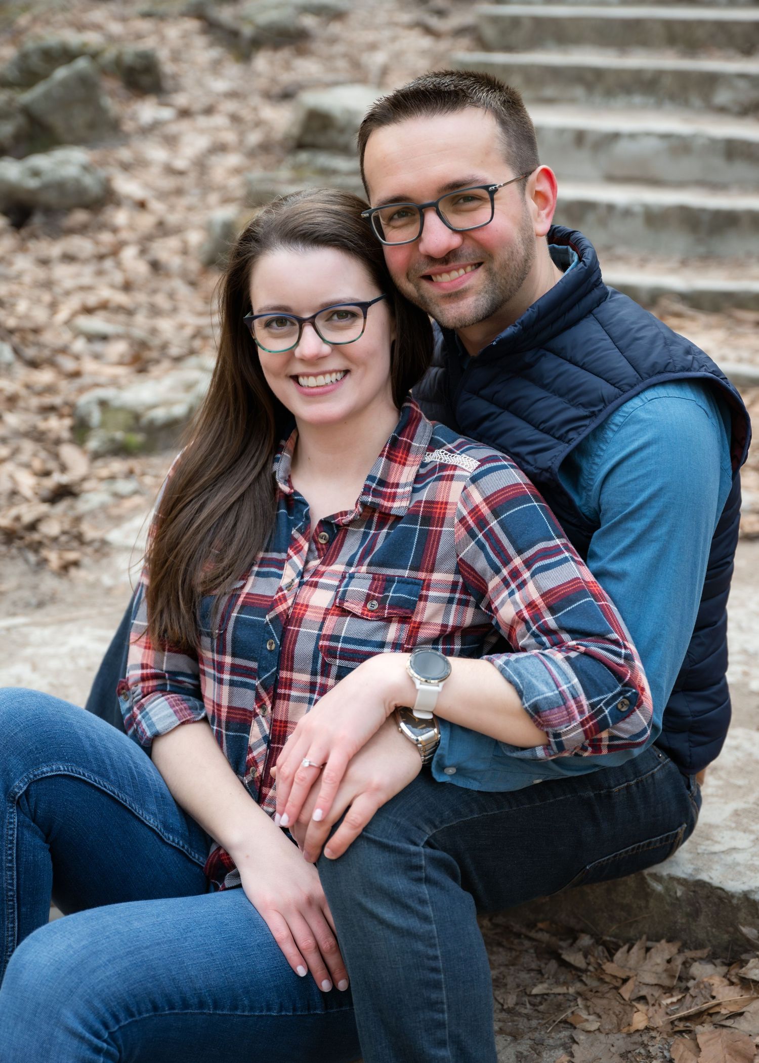 A smiling couple in plaid and navy sits on stone steps outdoors, surrounded by autumn leaves.