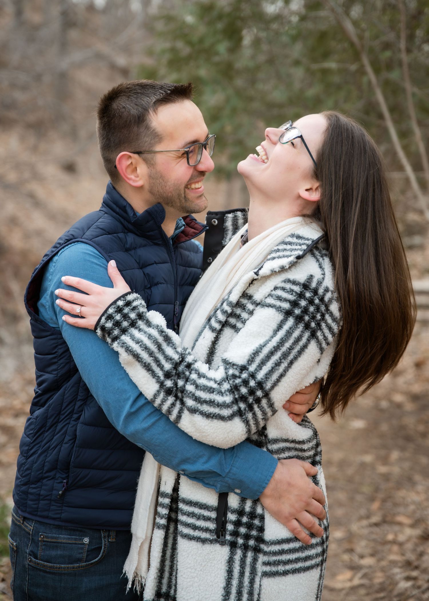 A smiling couple embraces outdoors in fall, wearing glasses and cozy layers, sharing a joyful moment on a wooded path.