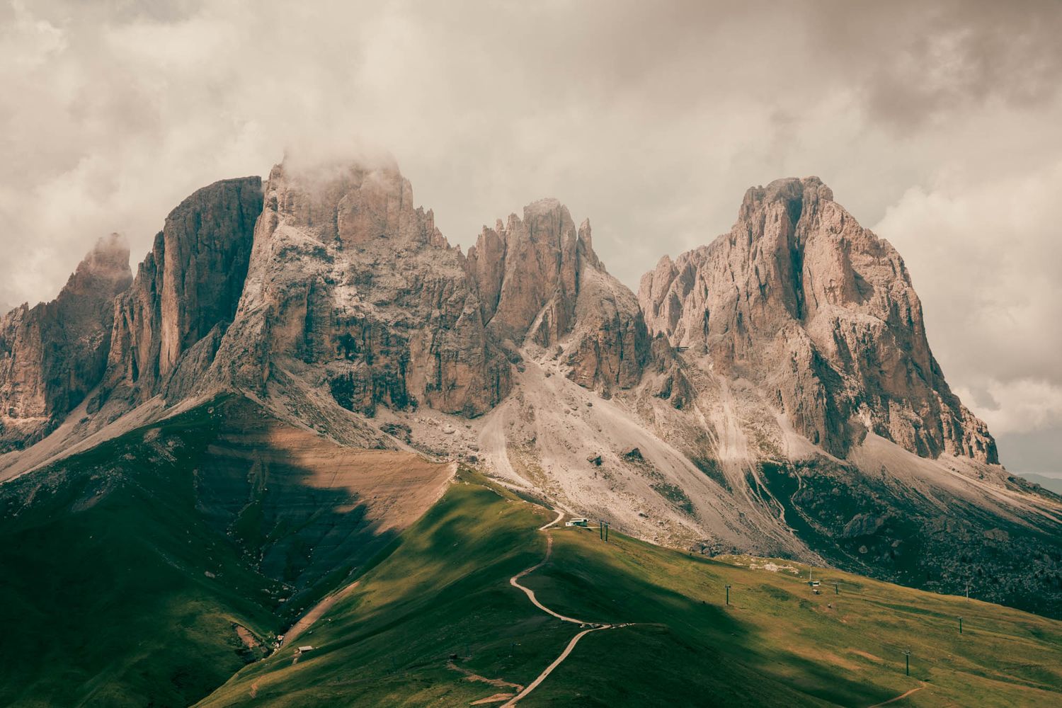 Dramatic rocky Dolomite peaks rise above green alpine meadows under a cloudy sky in northern Italy.