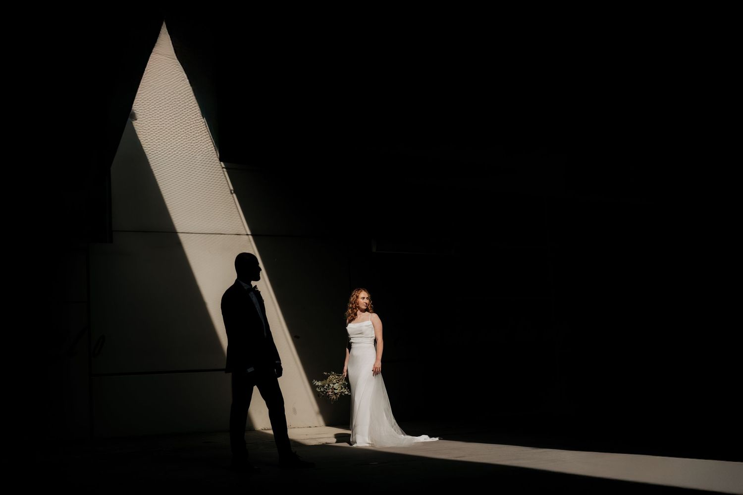 Bride in white gown and groom silhouette stand in dramatic light and shadow against a dark wall.