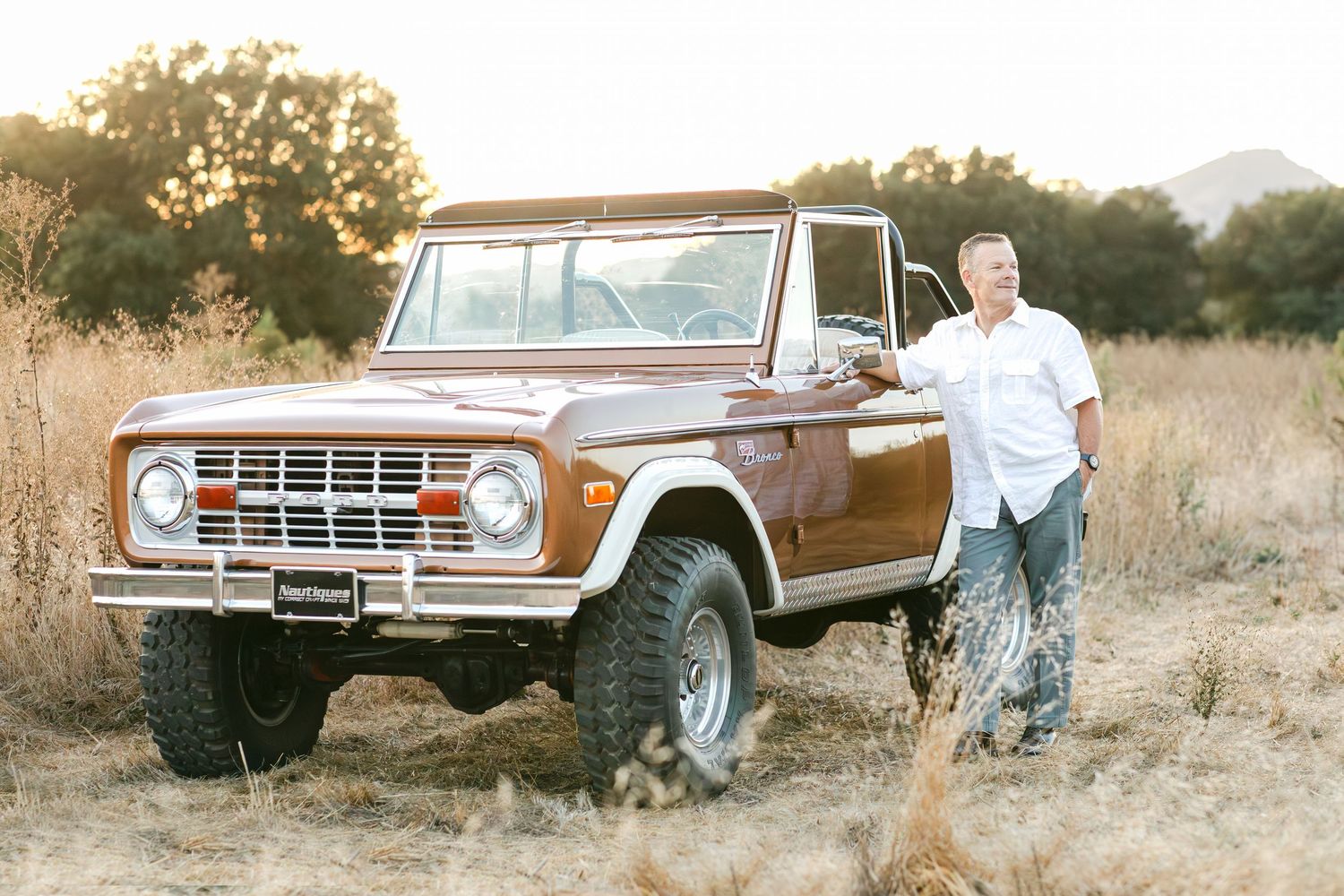 Man posing beside a classic restored Ford Bronco in a golden field during sunset.