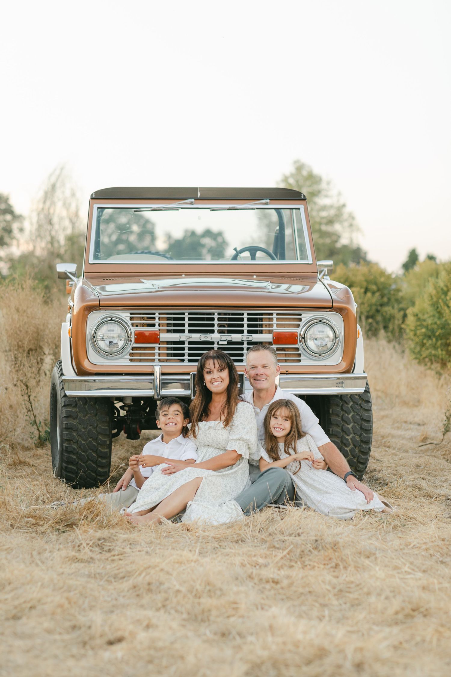 Family of four sitting in front of a vintage Ford Bronco in a golden field during a warm outdoor photo session.