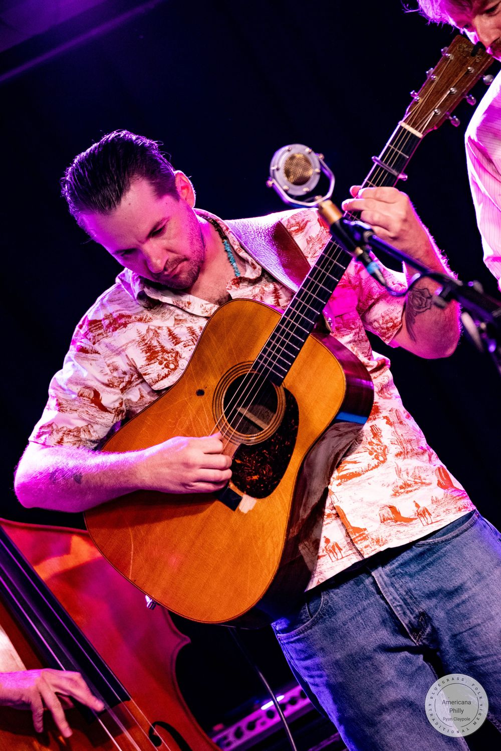 Musician playing an acoustic guitar on stage under warm purple stage lights at a live concert.