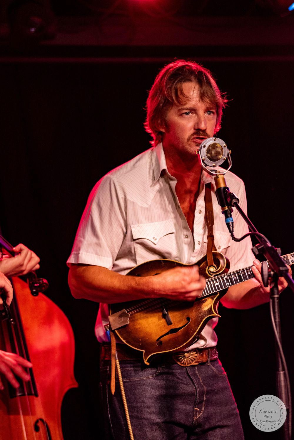 Male musician sings into vintage microphone while playing mandolin on stage under red stage lights.