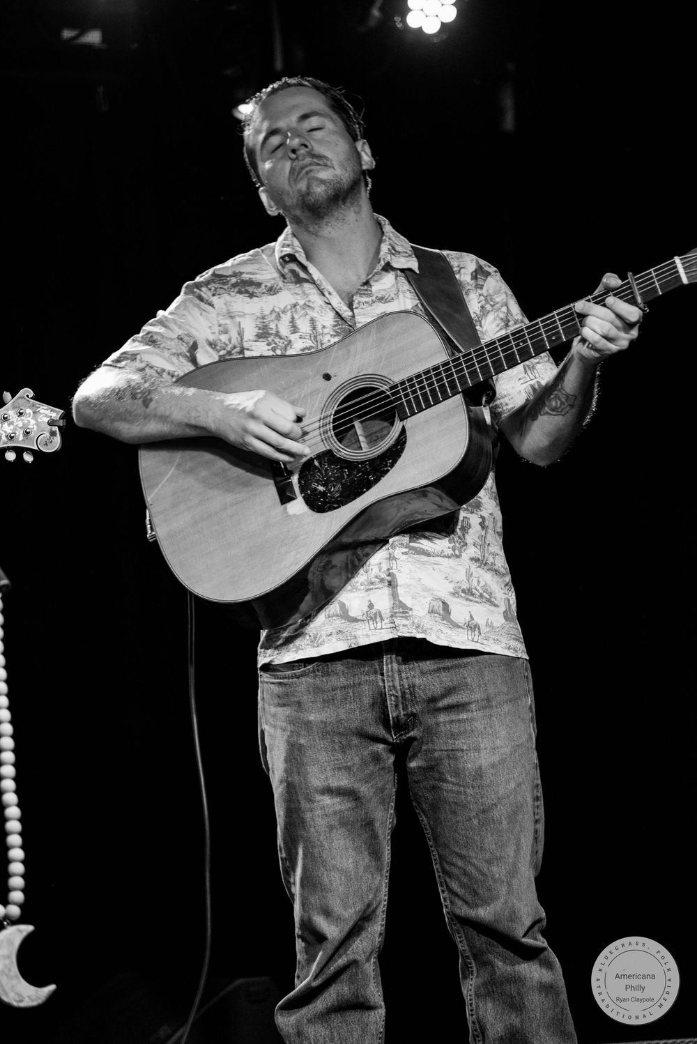 Black and white photo of male guitarist playing acoustic guitar with eyes closed on stage.