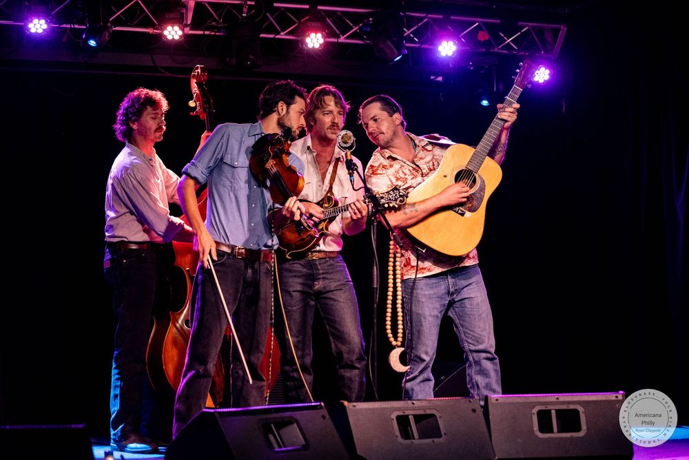 Bluegrass band of four male musicians performing together on stage sharing a microphone at a live show.