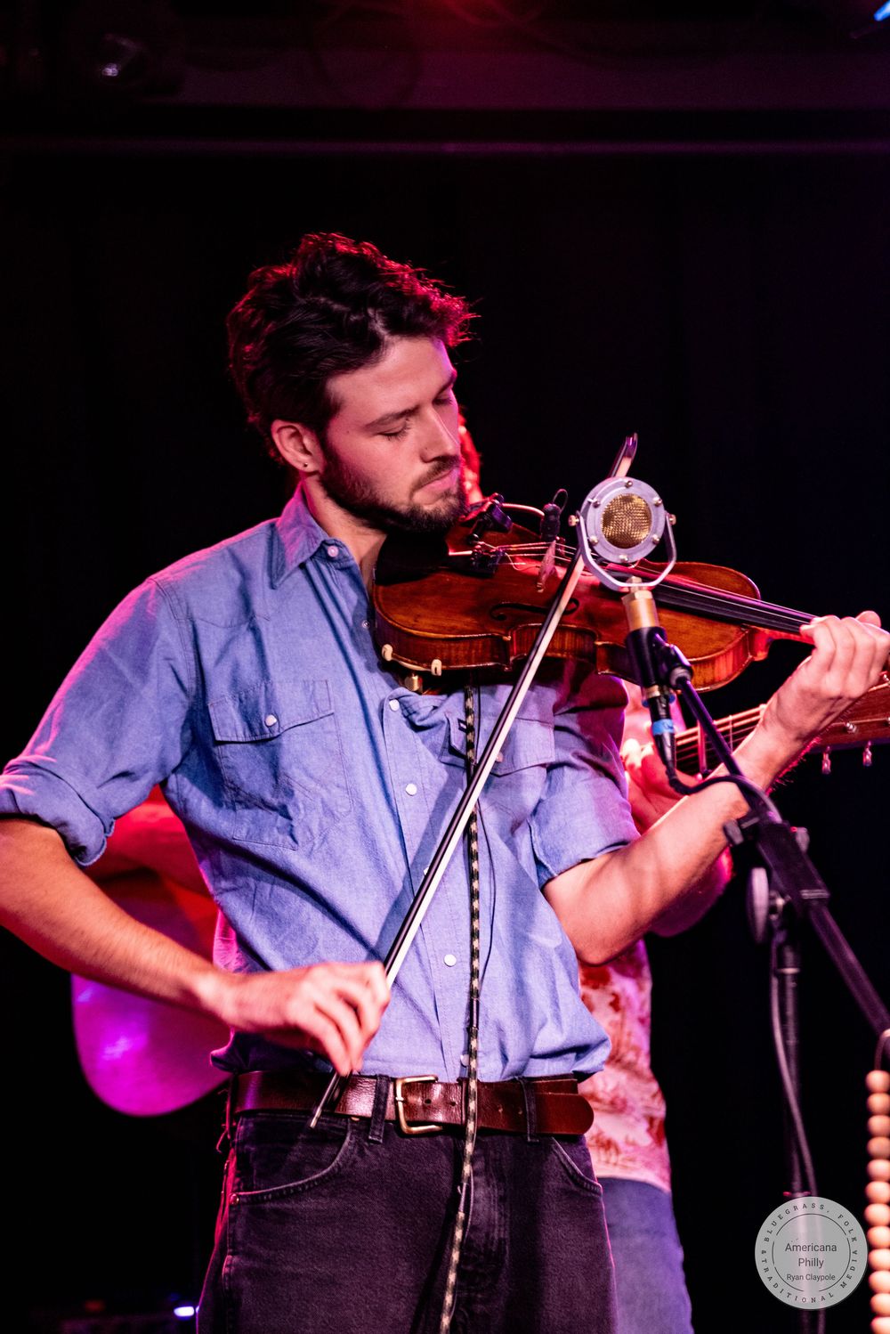 Bearded musician playing violin on stage in blue chambray shirt under dramatic red and blue stage lighting.