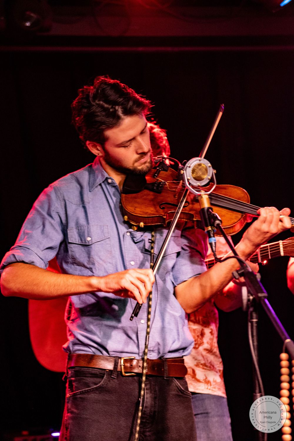 Bearded male violinist performs on stage under red and purple stage lighting at a live concert.