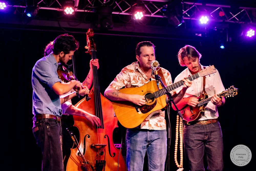 Three musicians perform bluegrass on stage, playing upright bass, acoustic guitar, and mandolin under purple lights.