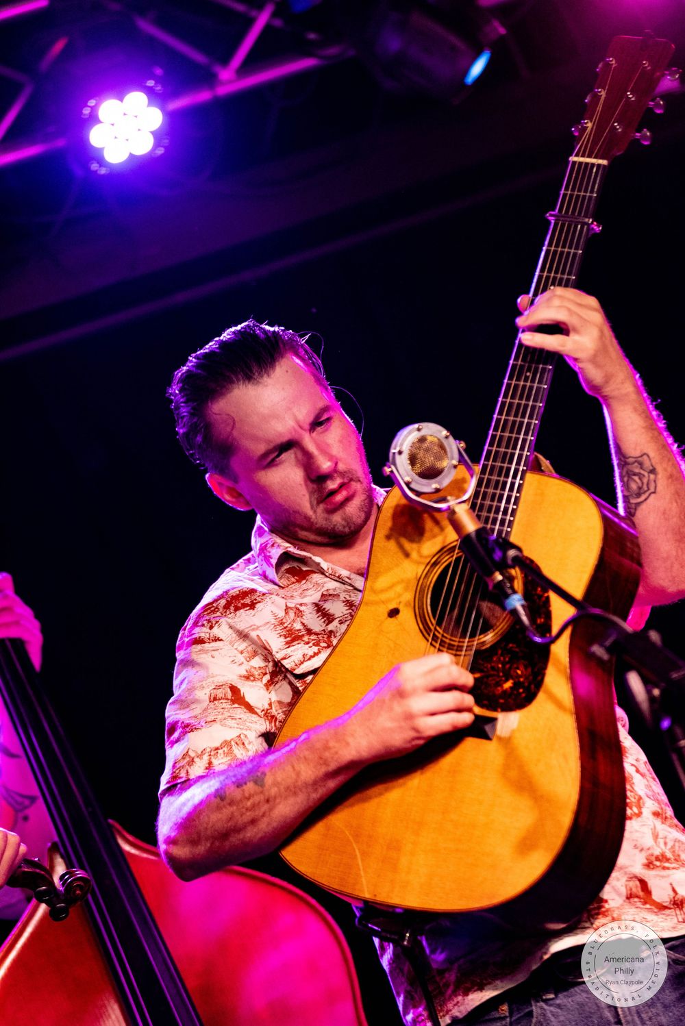 Male musician plays acoustic guitar on stage under purple and pink stage lights at a live concert.