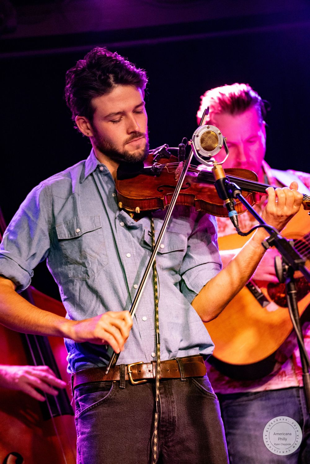 Young male violinist performing on stage under purple lights at a live bluegrass music concert.