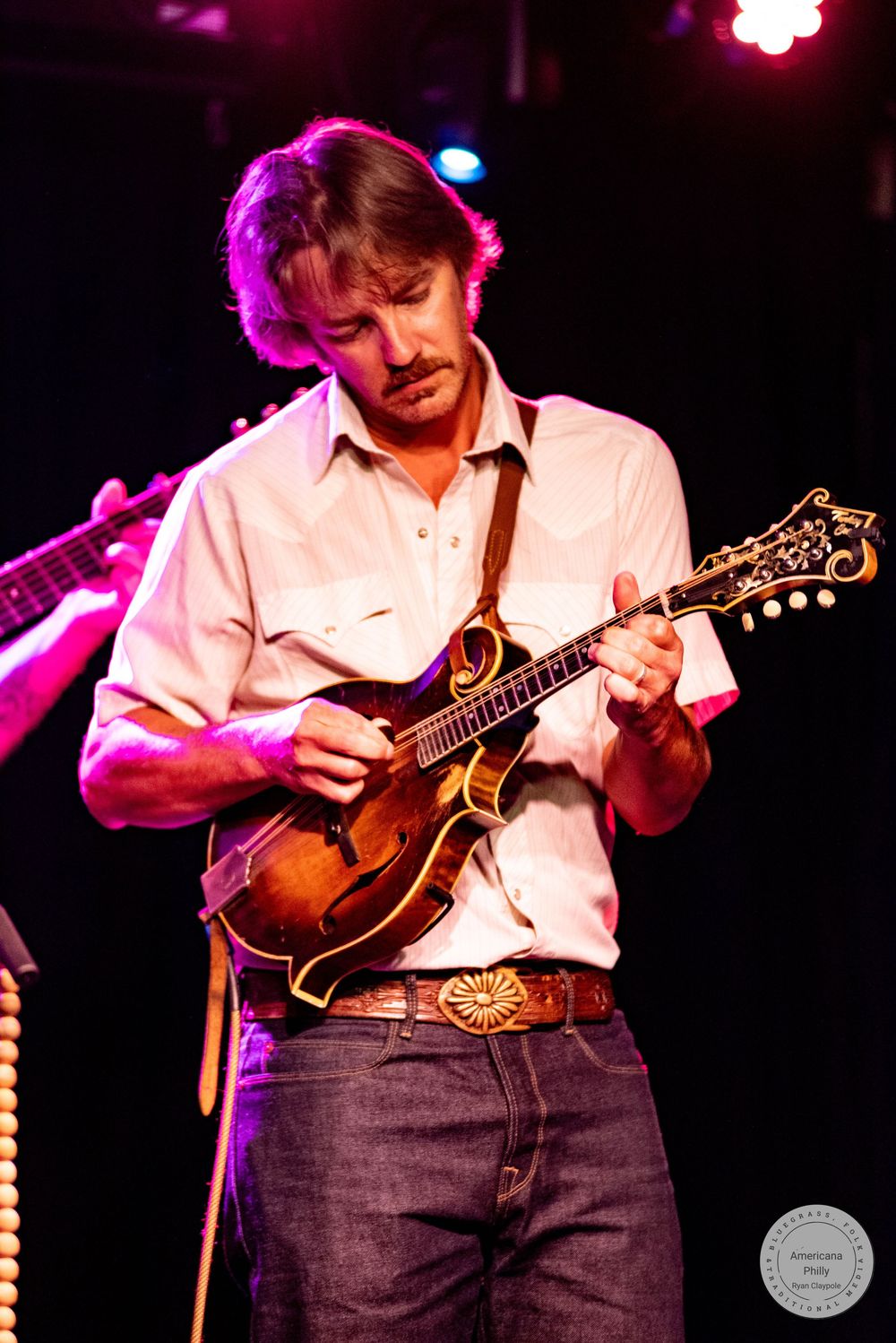 Man playing mandolin on stage in white shirt and jeans with decorative belt buckle under purple stage lighting.