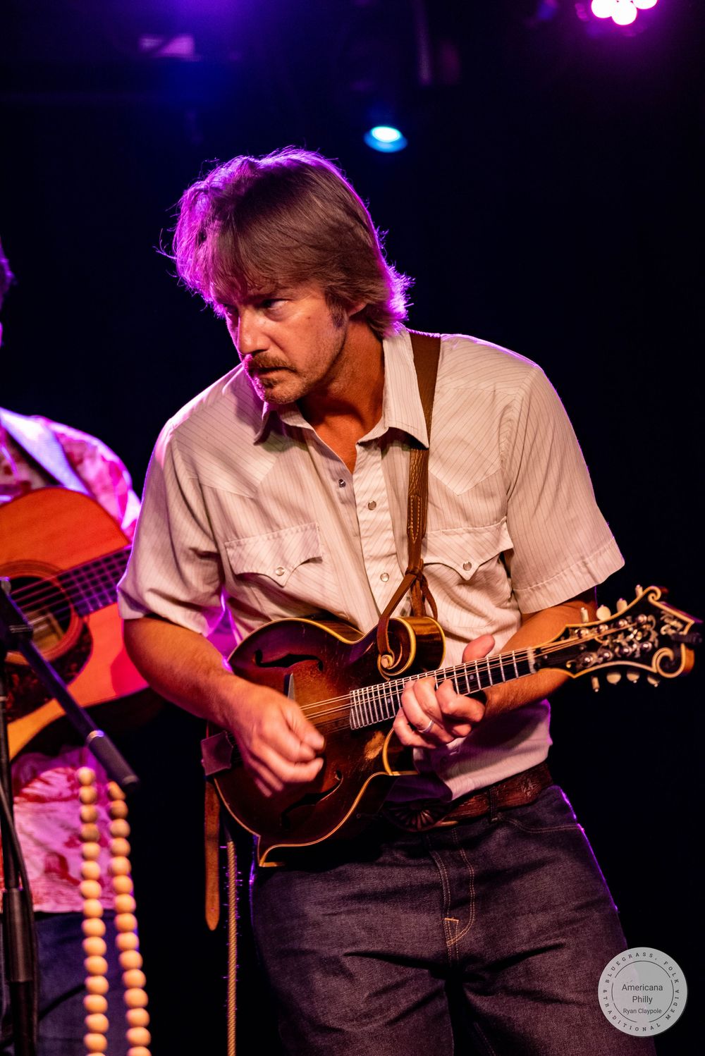 Male musician plays ornate mandolin on stage under purple lighting at a live bluegrass performance.