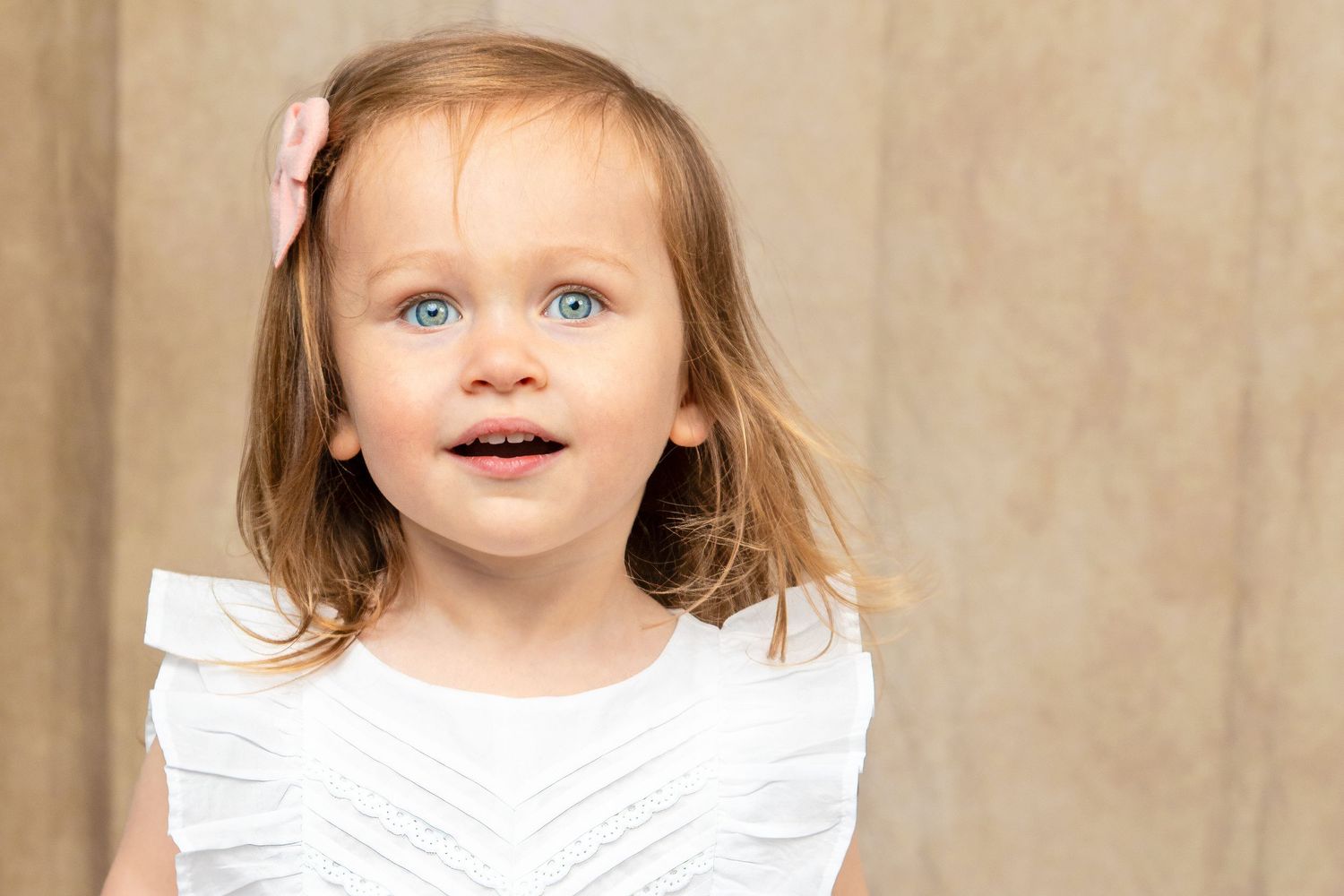 A cute toddler girl with blue eyes and a pink bow smiles in a white dress against a neutral background.