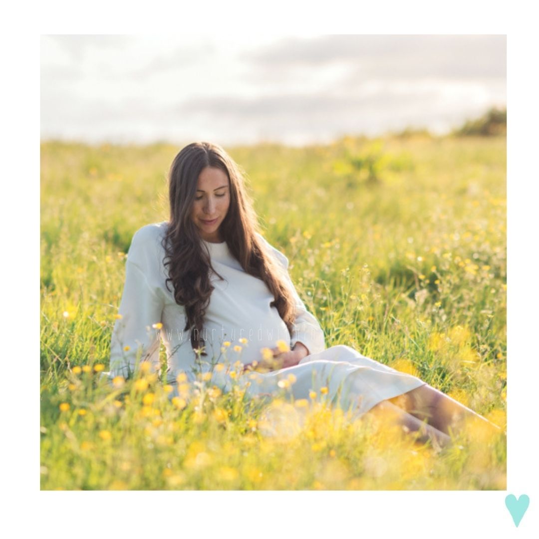 Pregnant woman in white dress sitting peacefully in a sunny field of yellow wildflowers, cradling her baby bump.