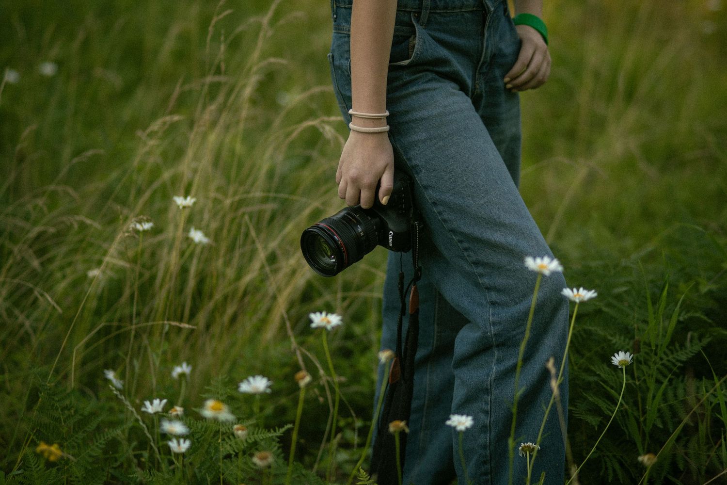 Person in jeans holding a DSLR camera standing in a lush green meadow with wildflowers.