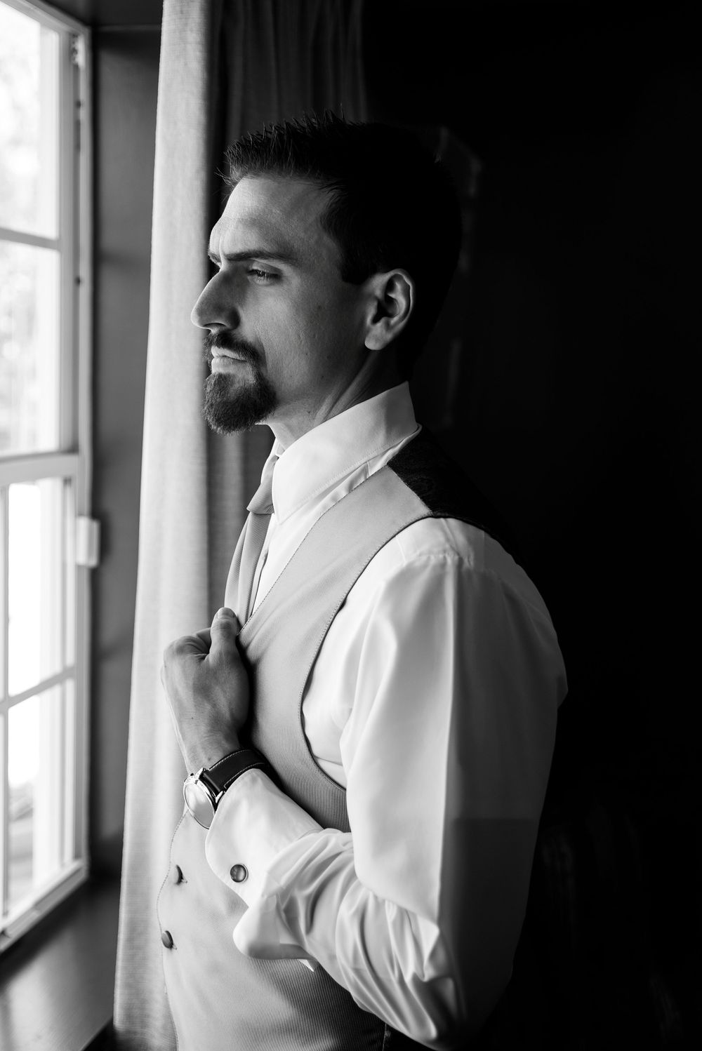Black and white photo of a well-dressed groom adjusting his tie by a window in a vest and dress shirt.