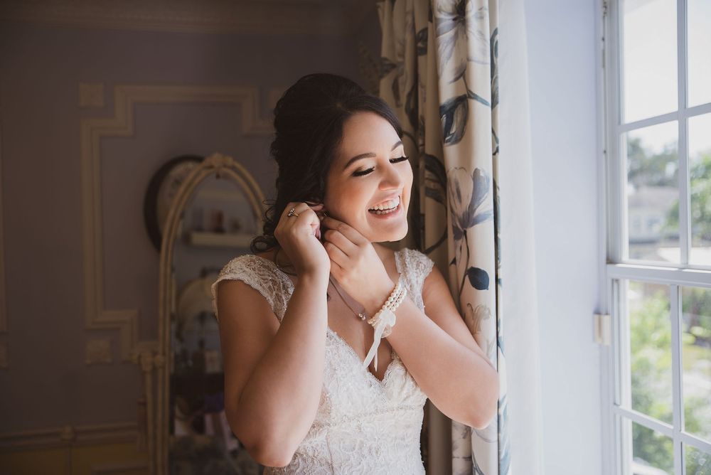 Bride smiling by window putting on earrings in white lace dress with elegant updo hairstyle and natural makeup.