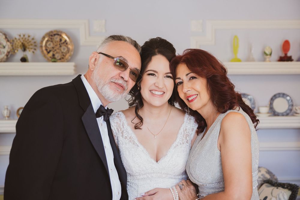 Bride smiling with parents on wedding day, woman in white lace gown flanked by father in tuxedo and mother in silver dress.