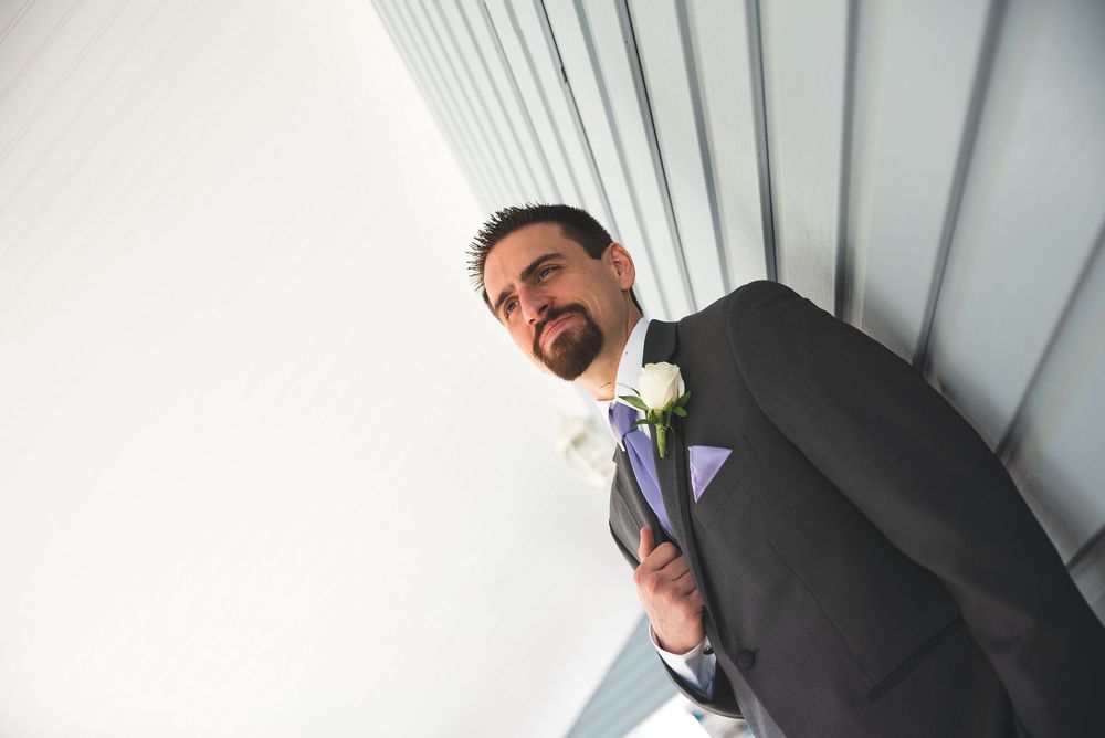 Groom in gray suit with white boutonniere and purple tie, photographed from below against white wall.