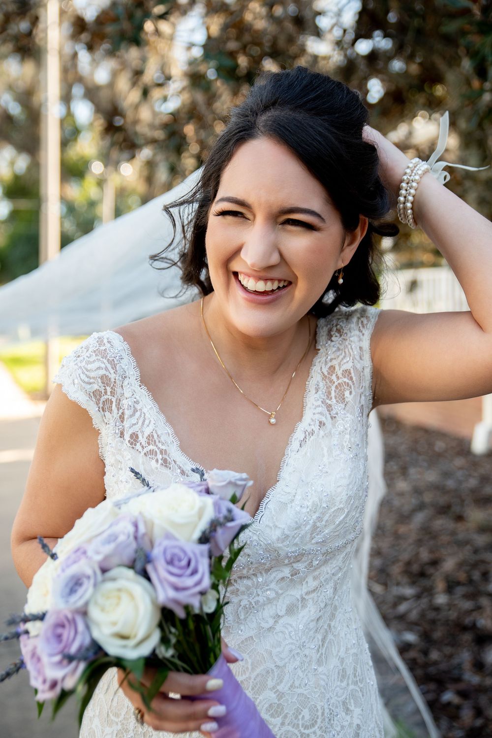 Smiling bride in lace wedding dress holding purple and white rose bouquet, veil blowing in the wind outdoors.
