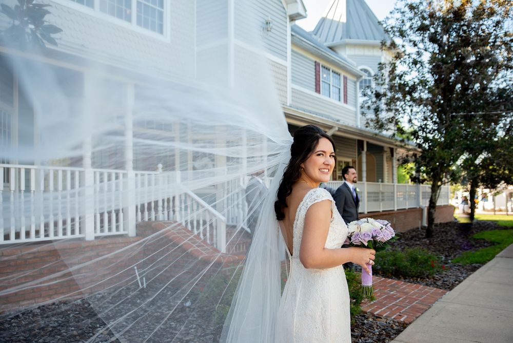 Bride in white gown with flowing veil caught by wind, holding bouquet, smiling outside elegant venue.