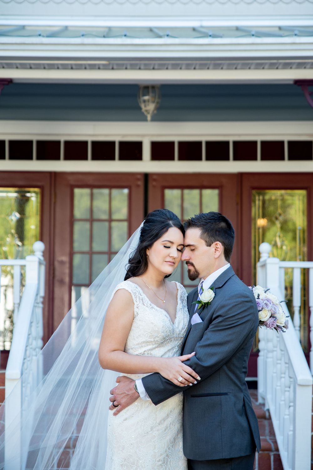 Bride and groom share a romantic forehead touch on wedding day, bride in lace gown with veil, groom in gray suit.