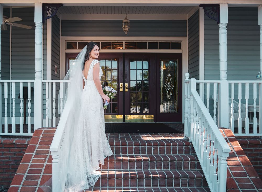 Bride in elegant white gown and veil poses on brick steps of charming Southern home with white porch railings.
