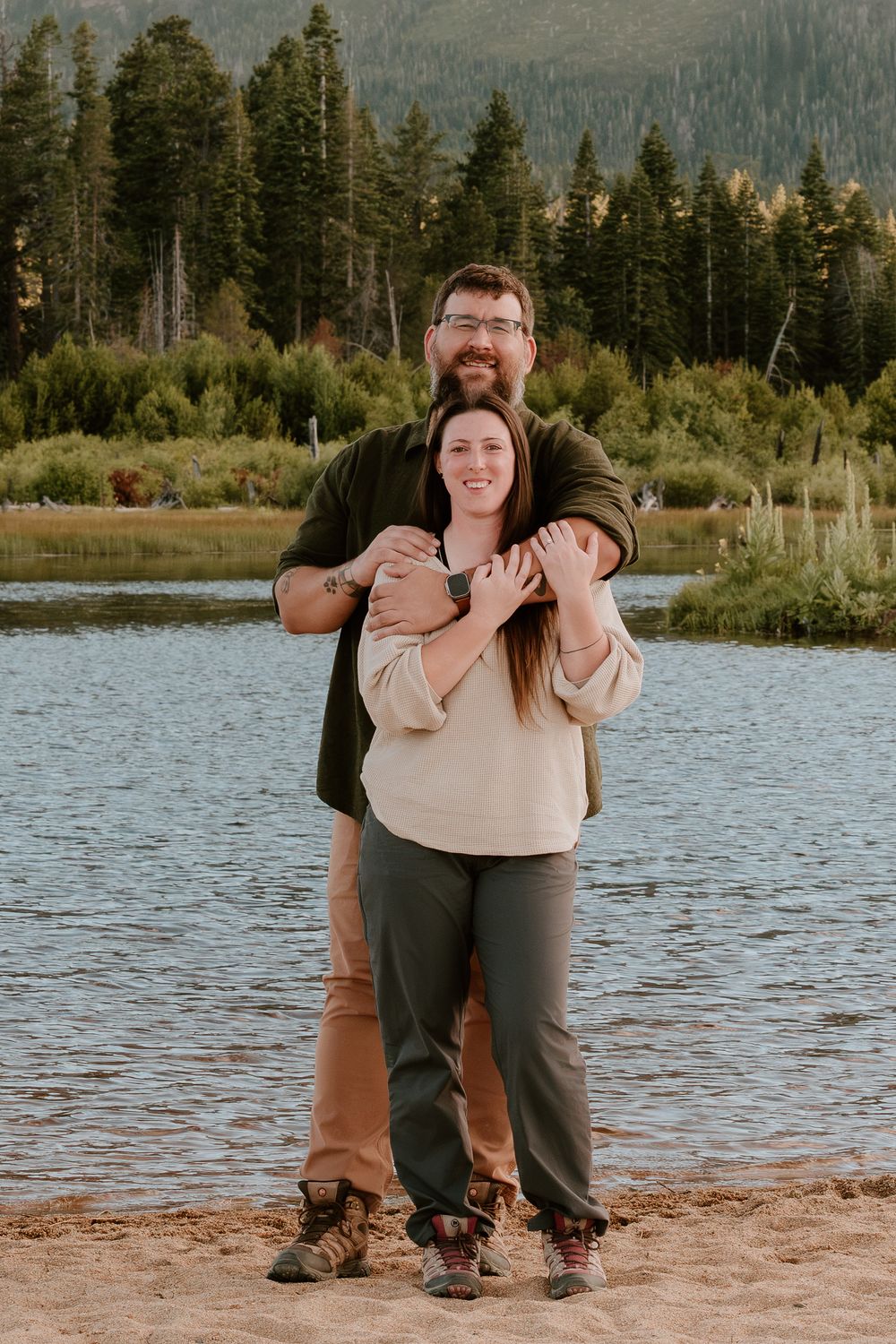 Couple embracing on a sandy lakeside beach surrounded by pine trees and mountains in a scenic outdoor portrait.
