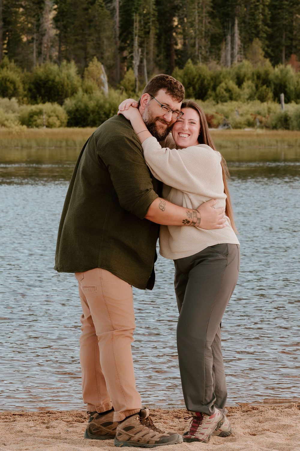 Happy couple hugging and smiling on a sandy lakeside beach surrounded by lush green trees.