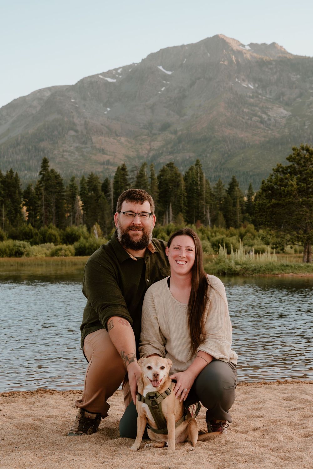 Couple and their yellow dog pose on sandy beach with mountain lake and pine trees backdrop at golden hour.